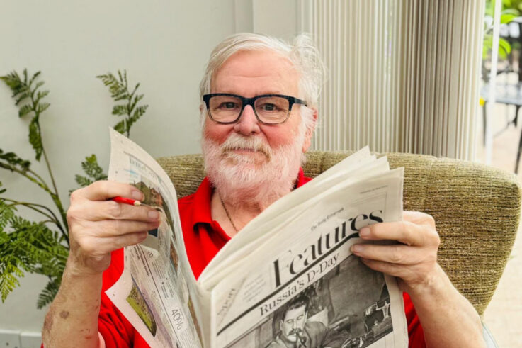 Older man with white hair and beard, wearing glasses and a red shirt, sits in a chair reading a newspaper titled Features. A plant and vertical blinds are visible in the background.