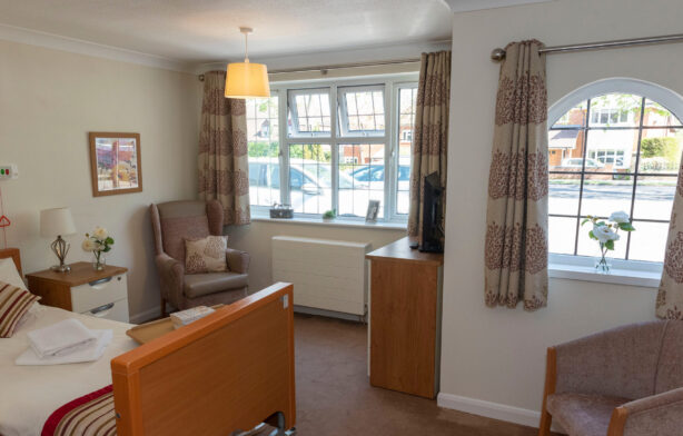 A neatly arranged bedroom with a single bed, armchair, small cupboard, window, and neutral-coloured curtains. Towels are folded on the bed.