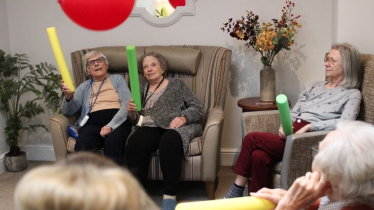 Three elderly women sit in armchairs, holding foam pool noodles and playing with a red balloon indoors. A vase with flowers is on a side table nearby.