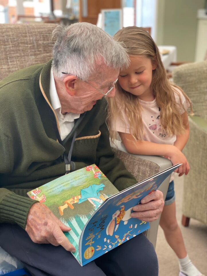 An elderly man reads a children's book to a young girl, who is smiling and leaning on his shoulder. They are seated indoors on upholstered chairs.