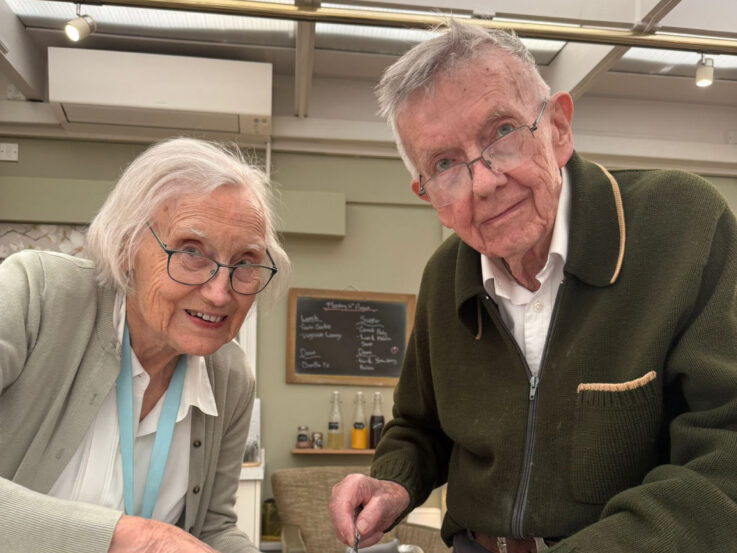 Two elderly people with glasses smile at the camera indoors, standing side by side in front of a chalkboard menu and drink bottles.