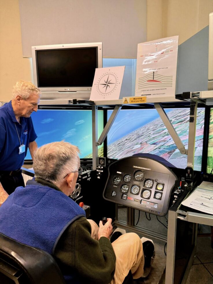 Two older men use a flight simulator with multiple monitors displaying flight data and a landscape, surrounded by instructional posters and aviation controls.