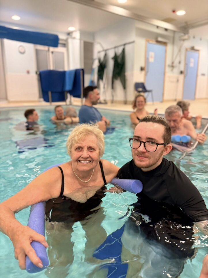 Group of adults exercising in an indoor swimming pool, holding foam noodles. An older woman and a younger man are at the front, smiling at the camera. Others are in the background.