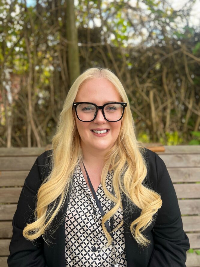 A woman with long blonde hair and glasses sits on a wooden bench outdoors, wearing a black blazer over a patterned blouse.