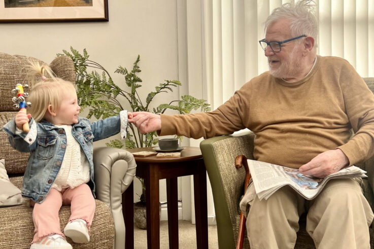 A toddler sitting on a chair hands a small object to an elderly man with a newspaper, as they both smile at each other in a lounge.