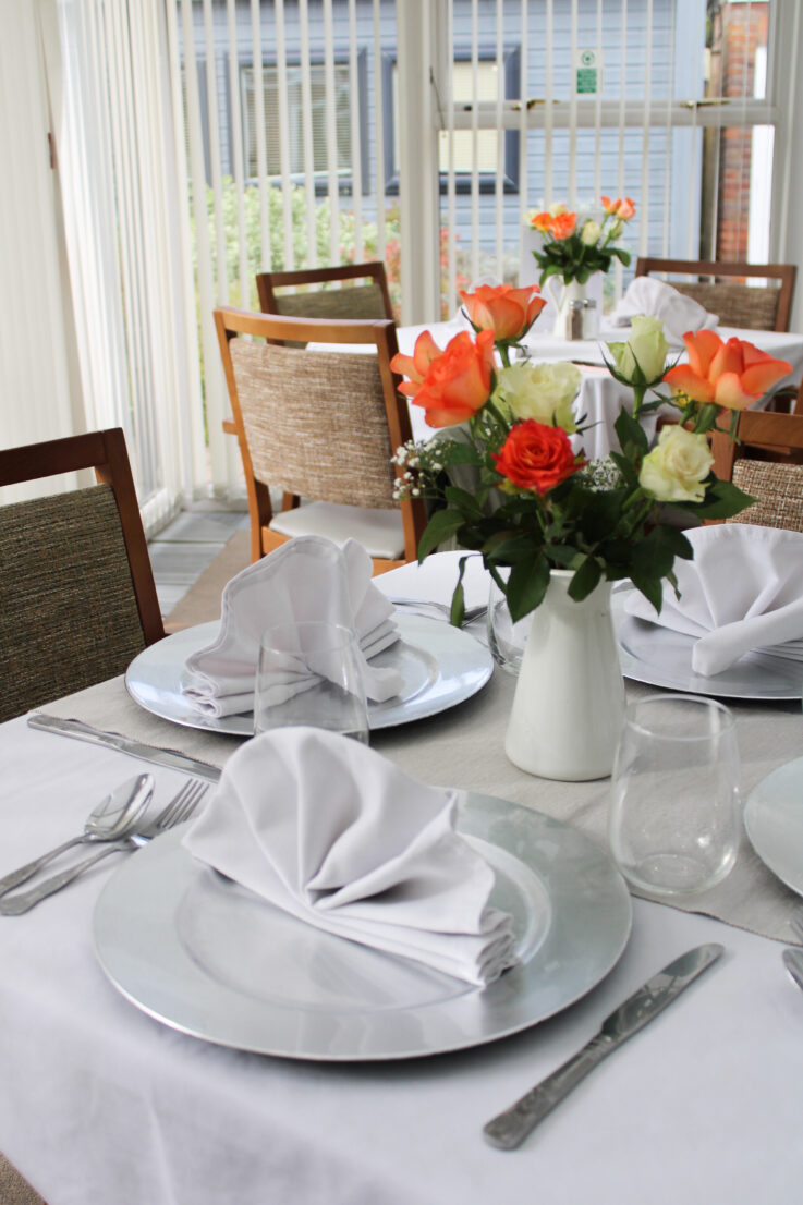 Table laid for a meal with white serviettes, silver plates, cutlery, glasses, and a vase of orange and white roses, in a bright room with large windows.