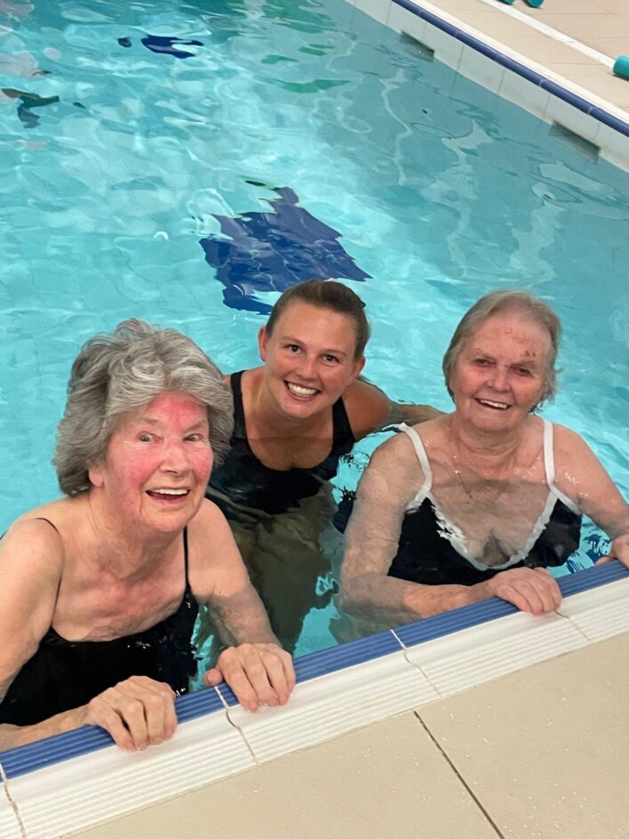 Three women in swimming costumes stand in a swimming pool, smiling at the camera and holding onto the pool edge.
