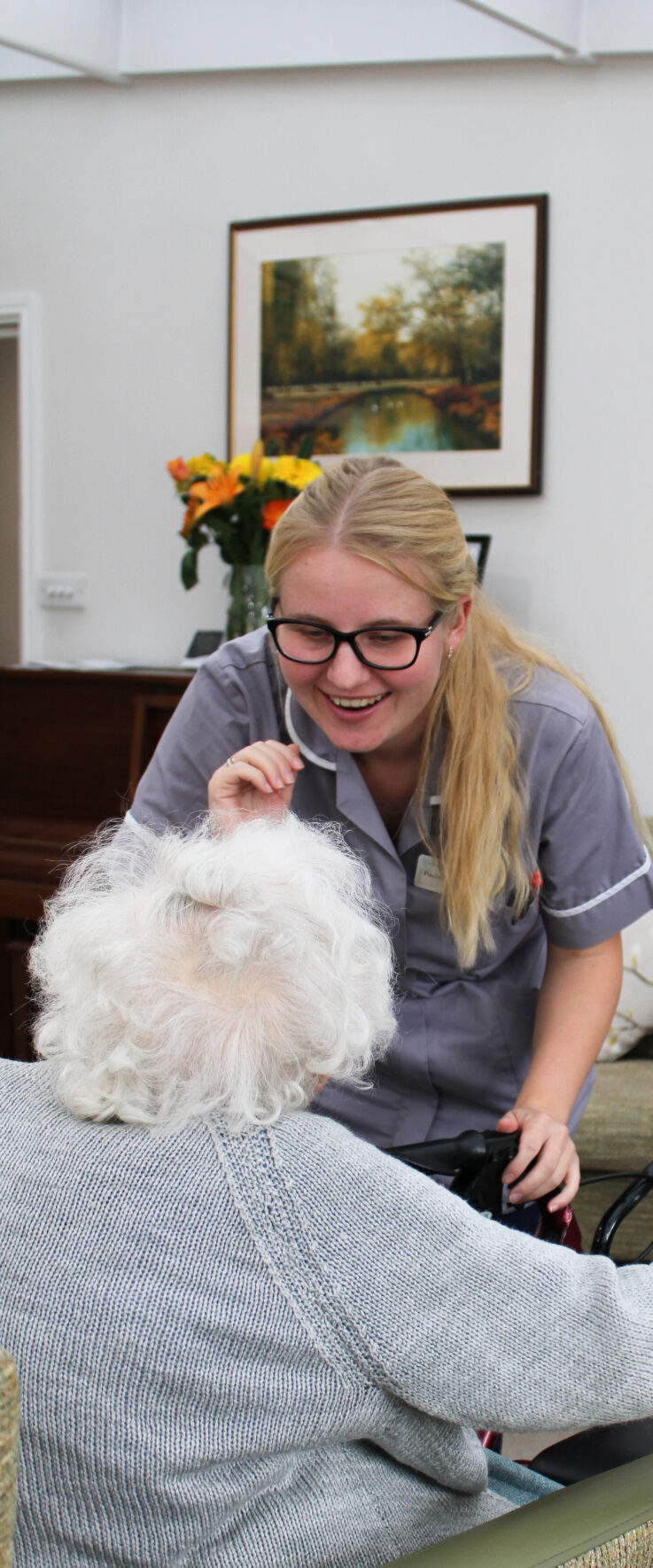 A carer in uniform smiles and speaks to an elderly person with white hair in a lounge with flowers and a landscape painting on the wall.