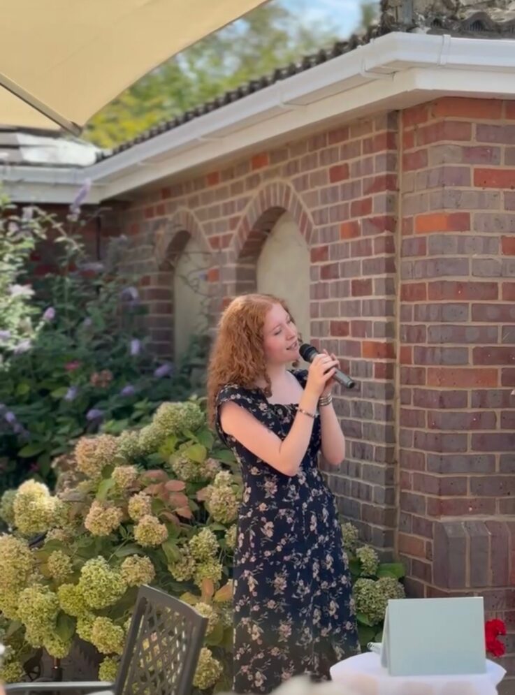 A person with curly red hair in a black floral dress sings into a microphone outdoors near a brick building and hydrangea bushes.