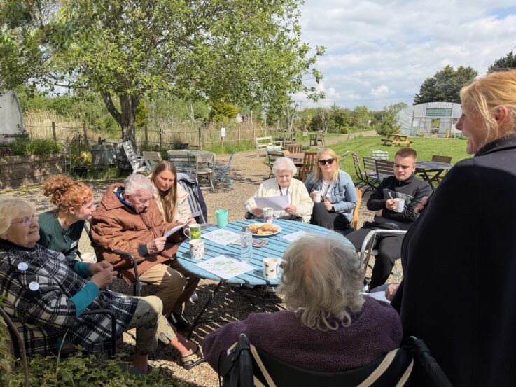 A group of people sit around a table outdoors, talking and reading papers. There are drinks and snacks on the table, and trees and garden structures in the background.