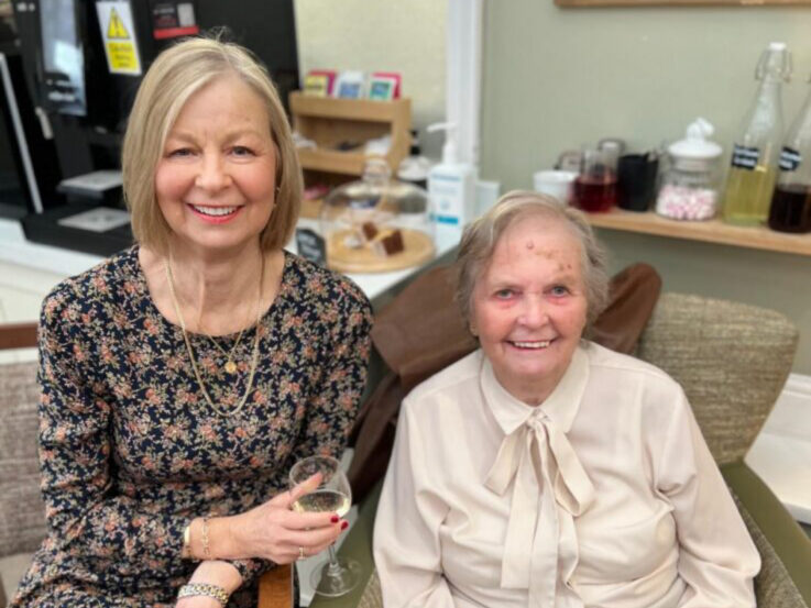 Two women sitting indoors, one holding a glass and smiling, the other in a cream blouse. Behind them are glass bottles, snacks, and kitchen items on shelves.