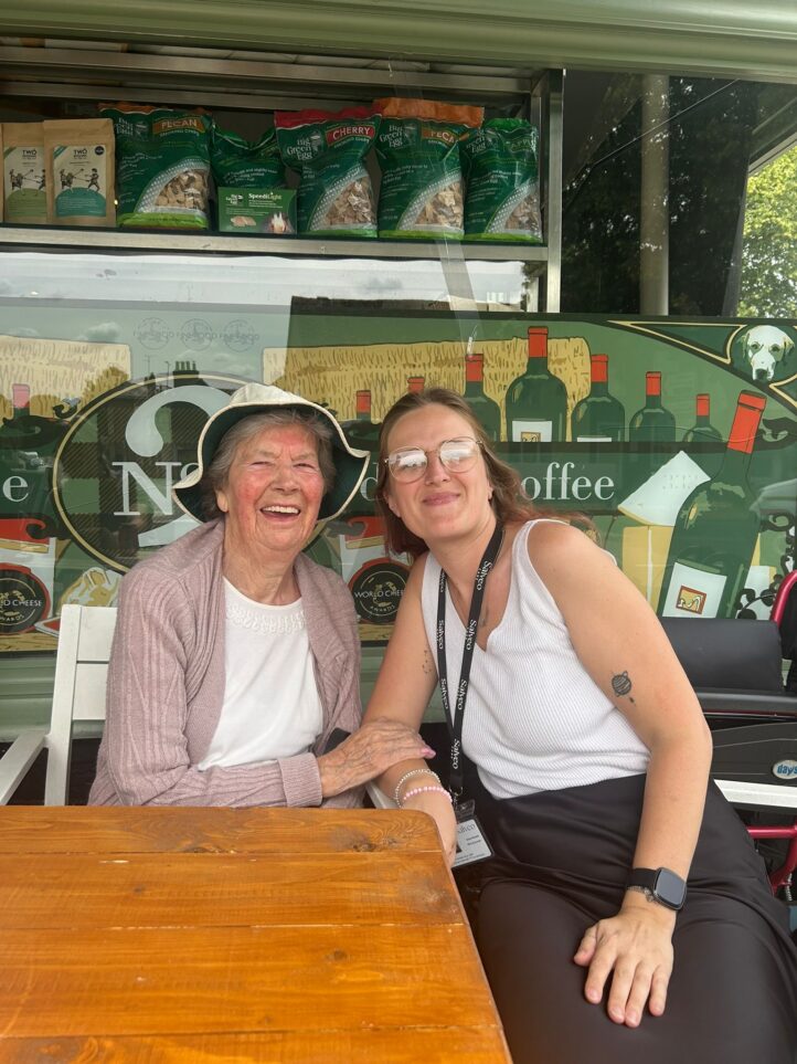 Two women sit together at a wooden table, smiling. Shelves with coffee and wine products are visible in the background.