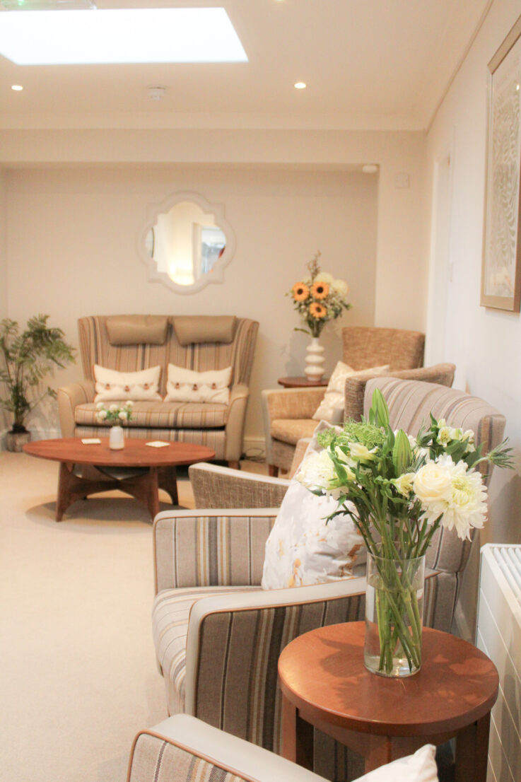 A bright, neutral-toned living room with striped armchairs, a sofa, coffee tables, flower arrangements, and decorative cushions. A skylight and wall mirror add light to the space.