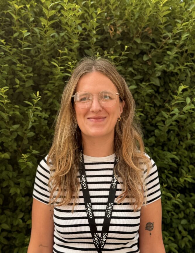 A person with long, light brown hair and glasses, wearing a striped shirt and a lanyard, stands in front of green foliage.