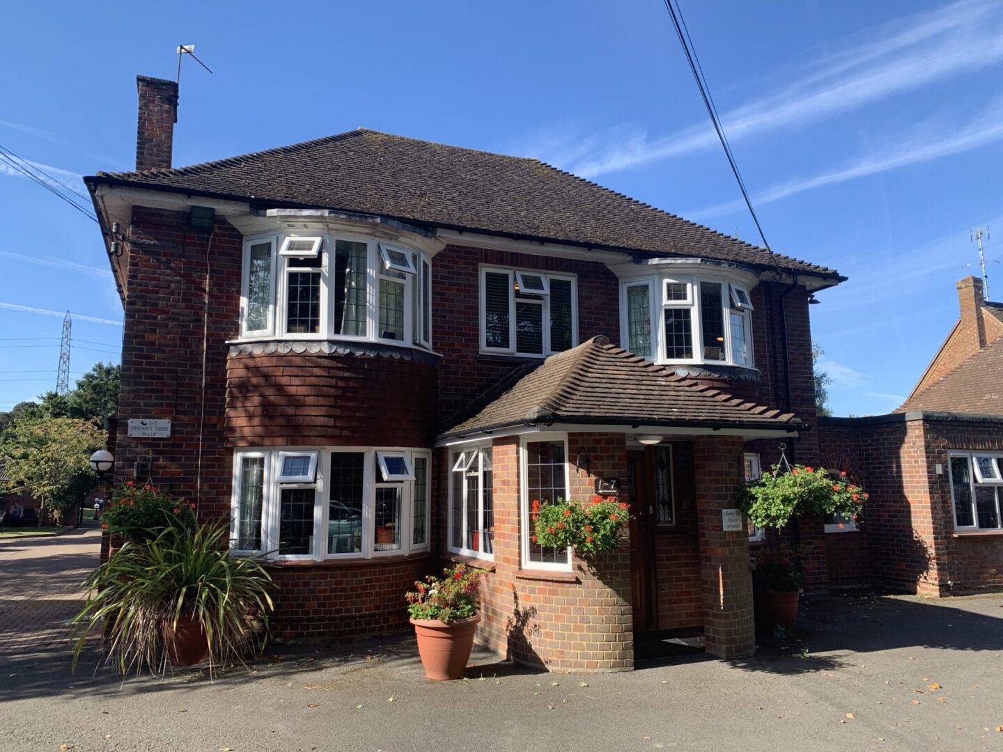 Two-storey red brick house with white trim, bay windows, and a small covered entrance. There are potted plants and hanging baskets near the front door.