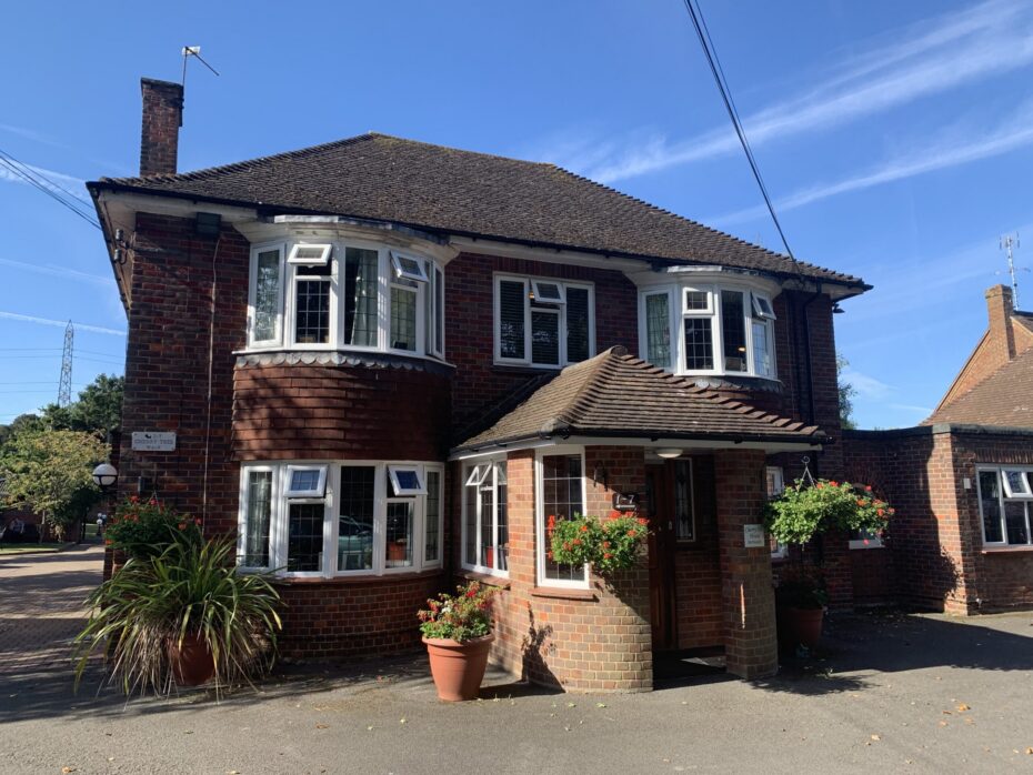 Two-storey red brick house with white trim, bay windows, and a small covered entrance. There are potted plants and hanging baskets near the front door.