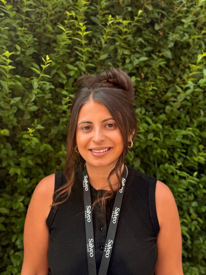 A person with long dark hair in a bun, wearing a sleeveless black top and a black lanyard, stands in front of green leafy shrubs.